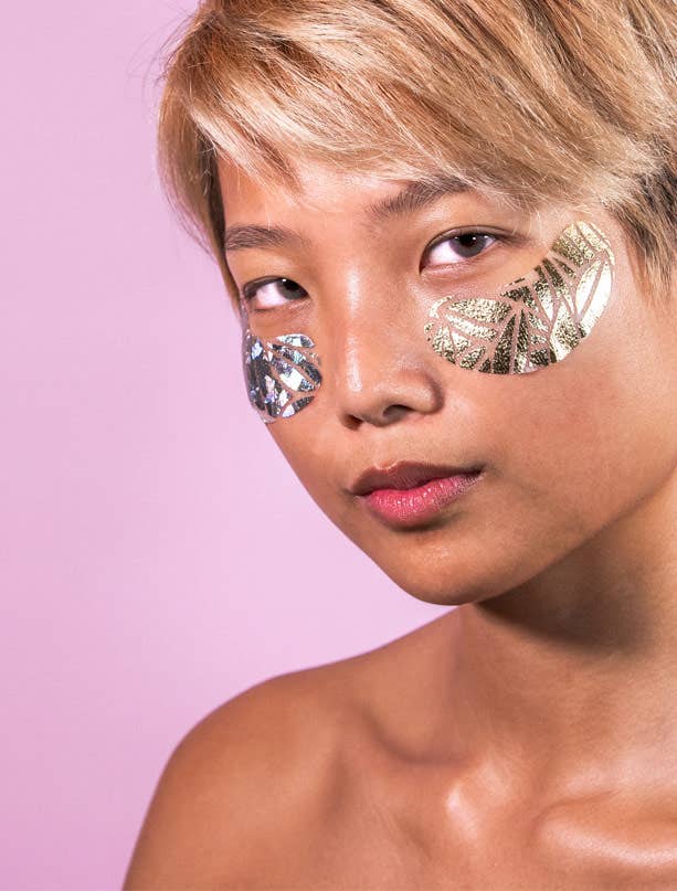 Woman wearing shimmering gold under-eye pads on bare skin against a pastel pink background — luxury skincare for brightening and depuffing tired eyes.