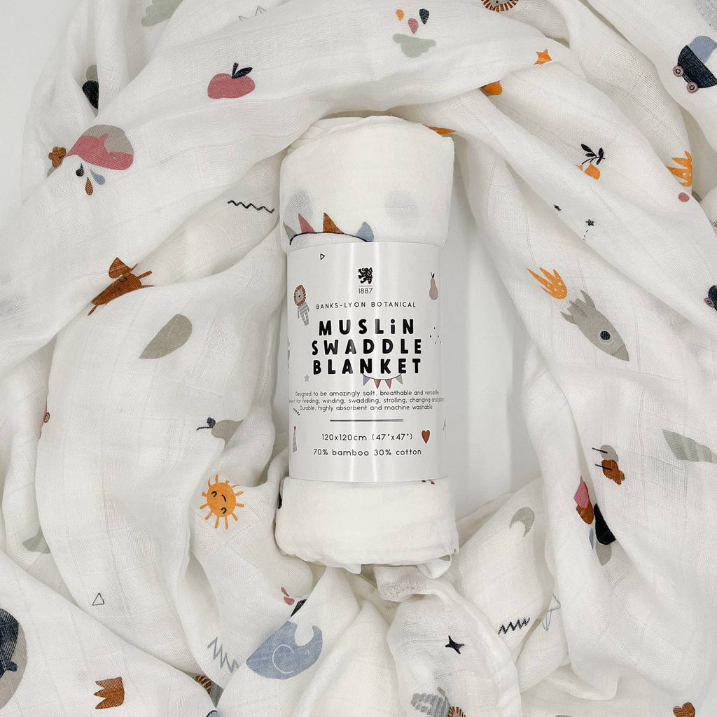 Soft organic muslin swaddle blanket by Banks Lyon Botanical, rolled and surrounded by matching fabric featuring whimsical illustrations of animals, fruit, rainbows, clouds, and suns.