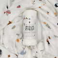 Soft organic muslin swaddle blanket by Banks Lyon Botanical, rolled and surrounded by matching fabric featuring whimsical illustrations of animals, fruit, rainbows, clouds, and suns.