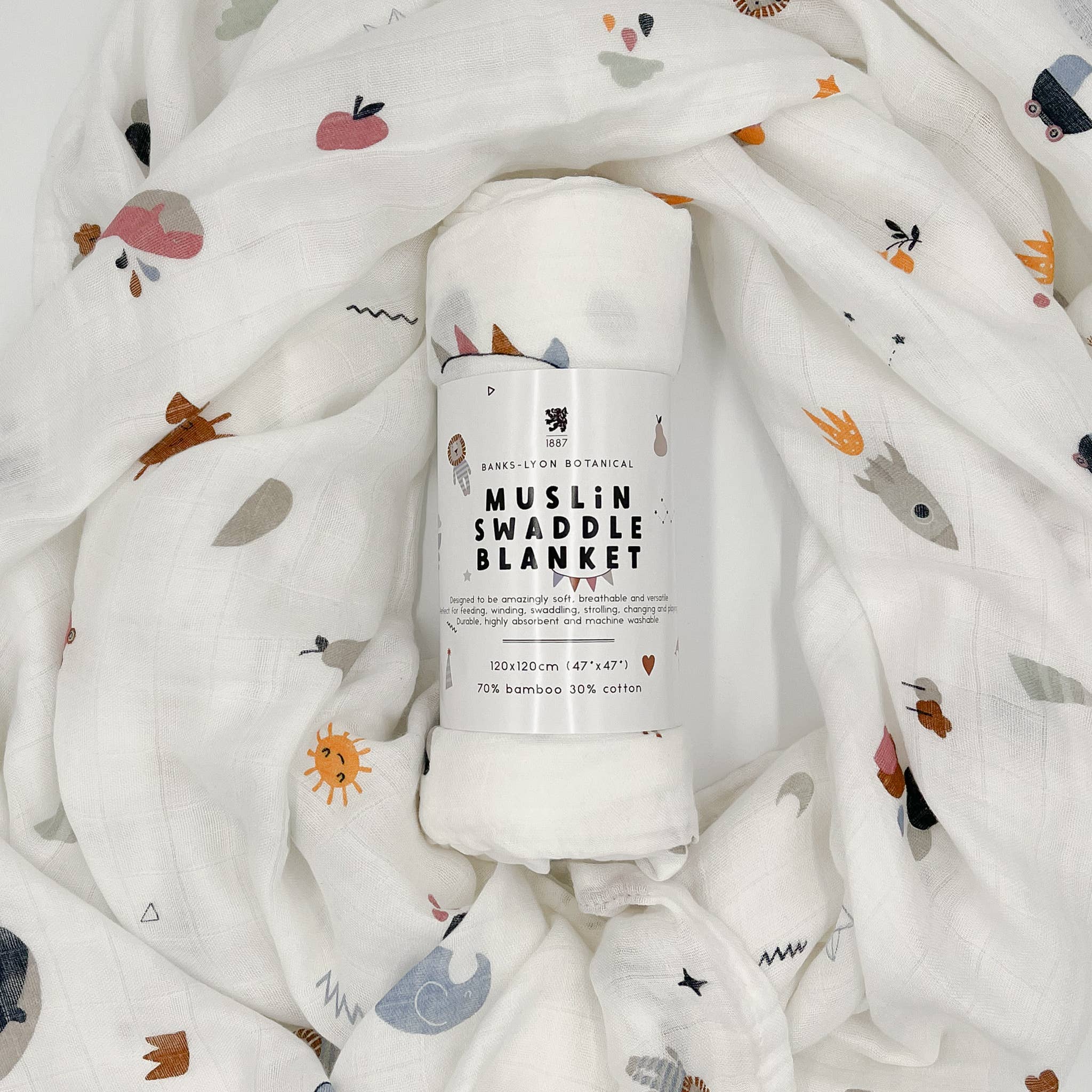 Soft organic muslin swaddle blanket by Banks Lyon Botanical, rolled and surrounded by matching fabric featuring whimsical illustrations of animals, fruit, rainbows, clouds, and suns.