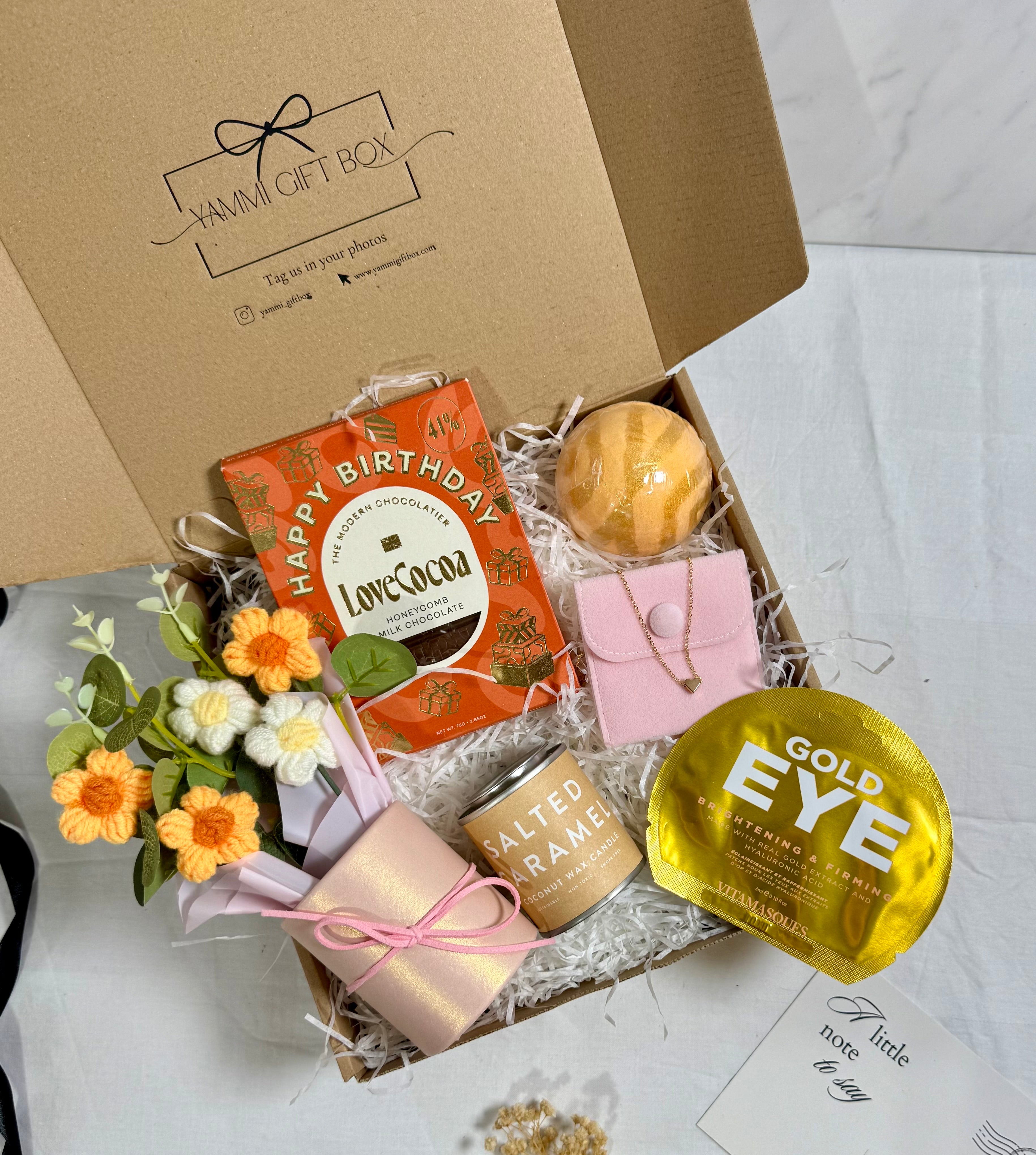 Gift box with various items including a birthday card, candle, and small plant on a white background.