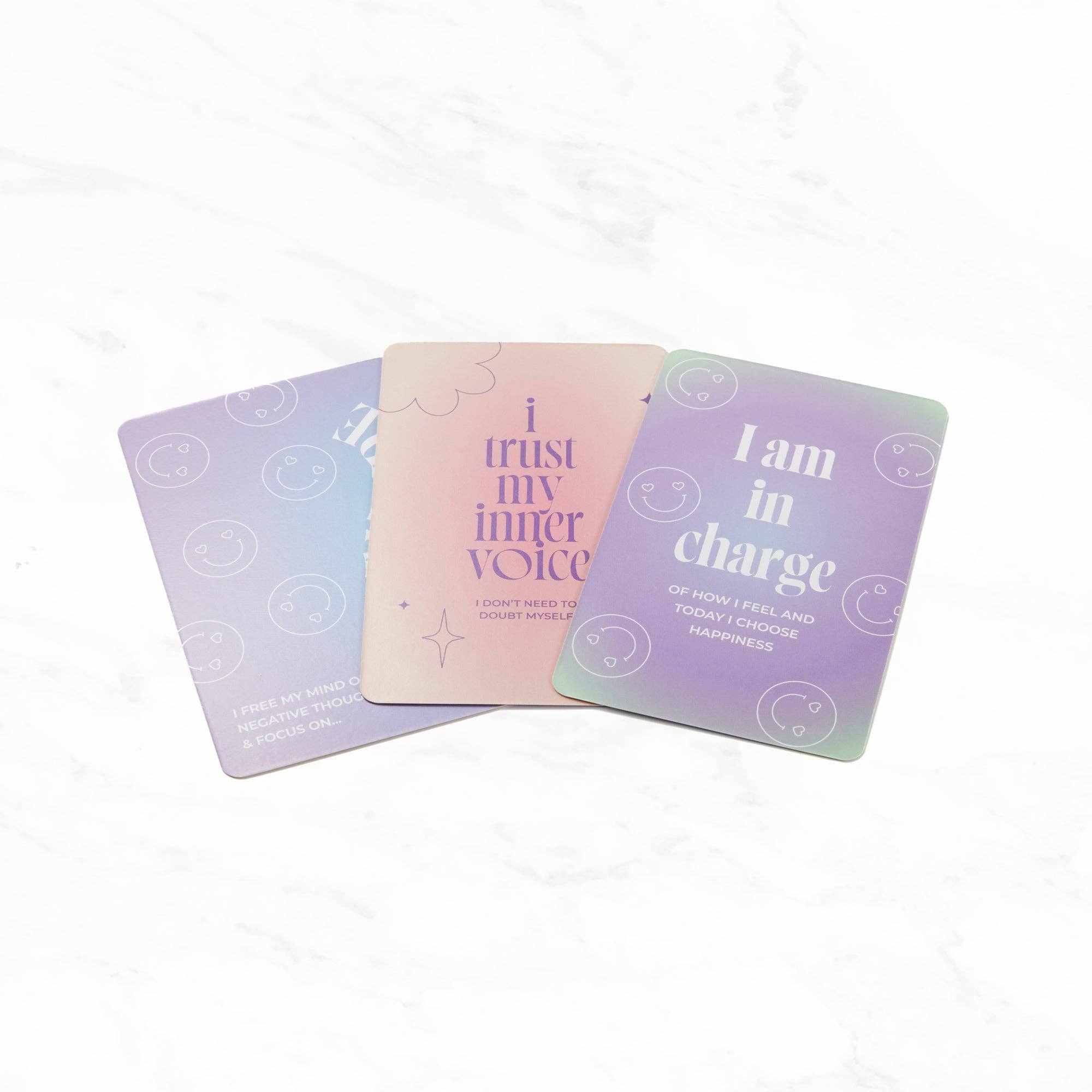 Three pastel gradient affirmation cards displayed on a white marble background. Each card features uplifting text with playful smiley face icons: one reads “I trust my inner voice,” another says “I am in charge of how I feel and today I choose happiness,” and the third states “I free my mind of negative thoughts & focus on...”. The cards have a soft, calming colour palette with tones of lilac, blush pink, and mint green, designed to inspire positivity and mindfulness.