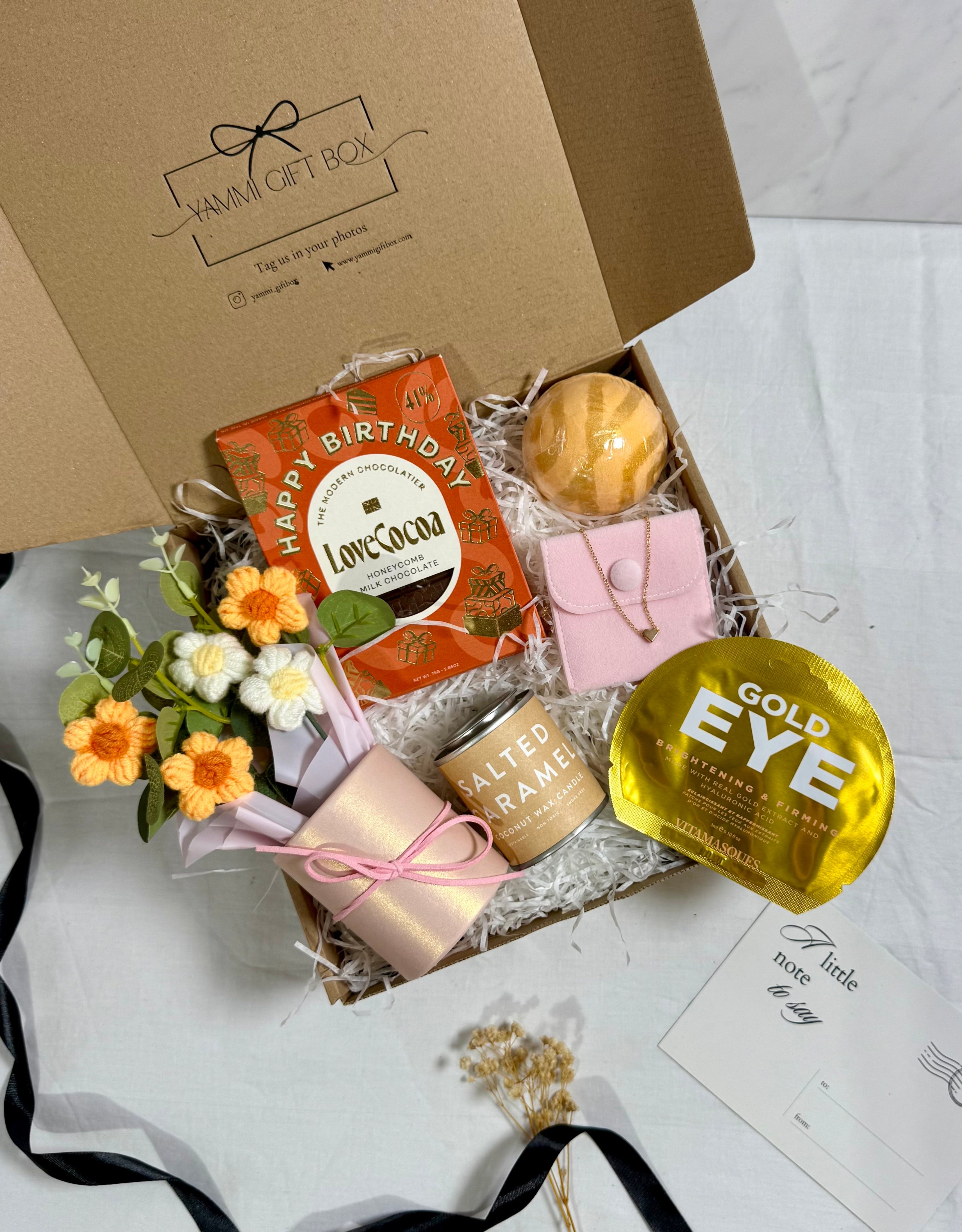 Gift box with various items including a mug, flowers, and a chocolate bar on a white background.