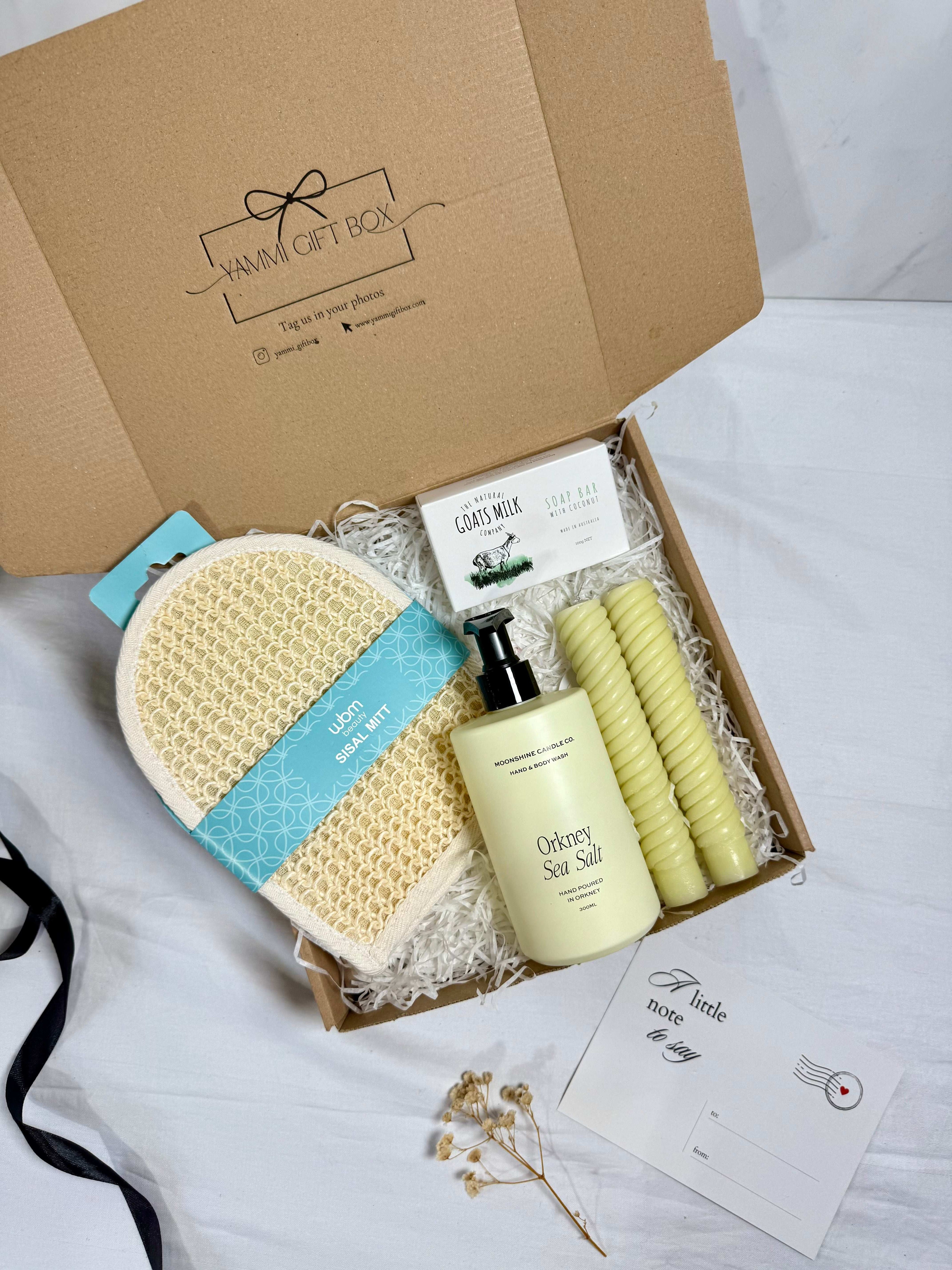 Gift box with bath products including a loofah, soap, and candles on a white surface.