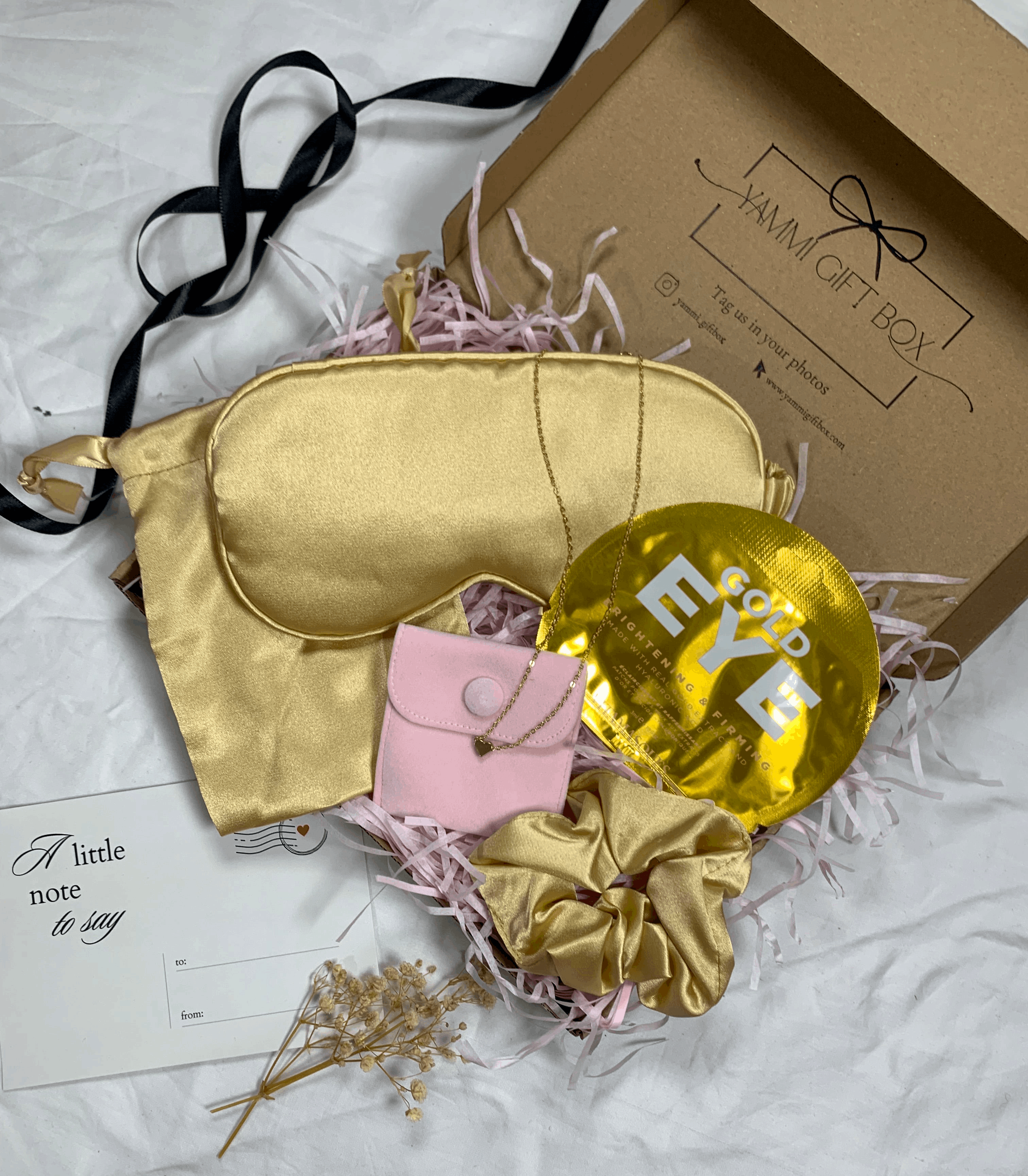 Luxury gold pamper gift box with satin eye mask, gold scrunchie, gold under-eye mask, pink pouch with delicate gold heart bracelet, dried flowers, and note card.