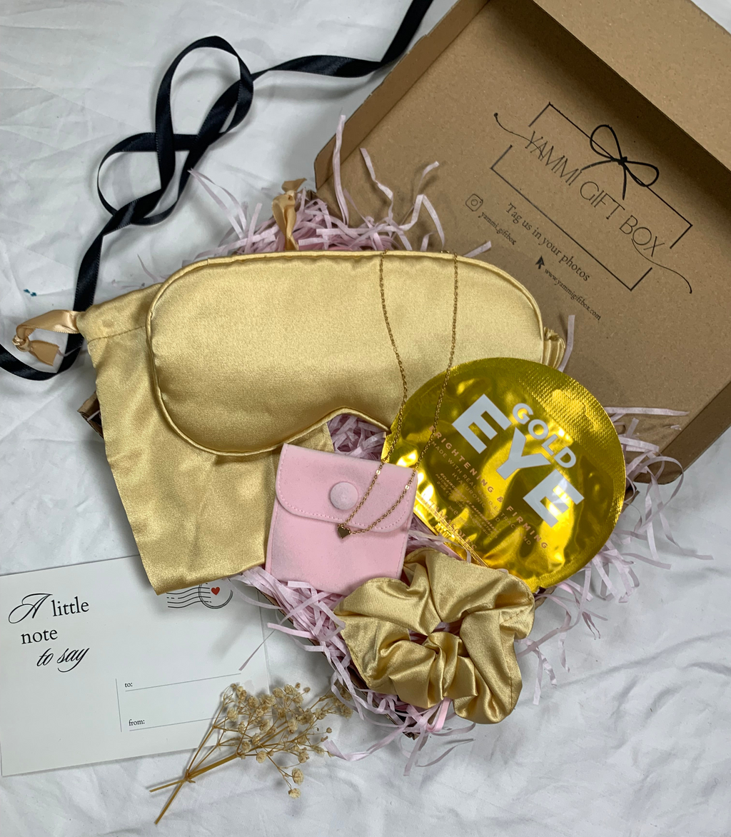 Luxury gold pamper gift box with satin eye mask, gold scrunchie, gold under-eye mask, pink pouch with delicate gold heart bracelet, dried flowers, and note card.