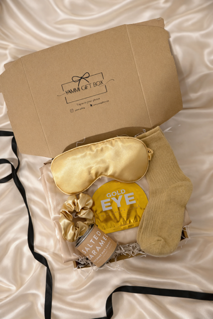 Gift box with gold sleep mask, socks, and other items on a white satin surface.