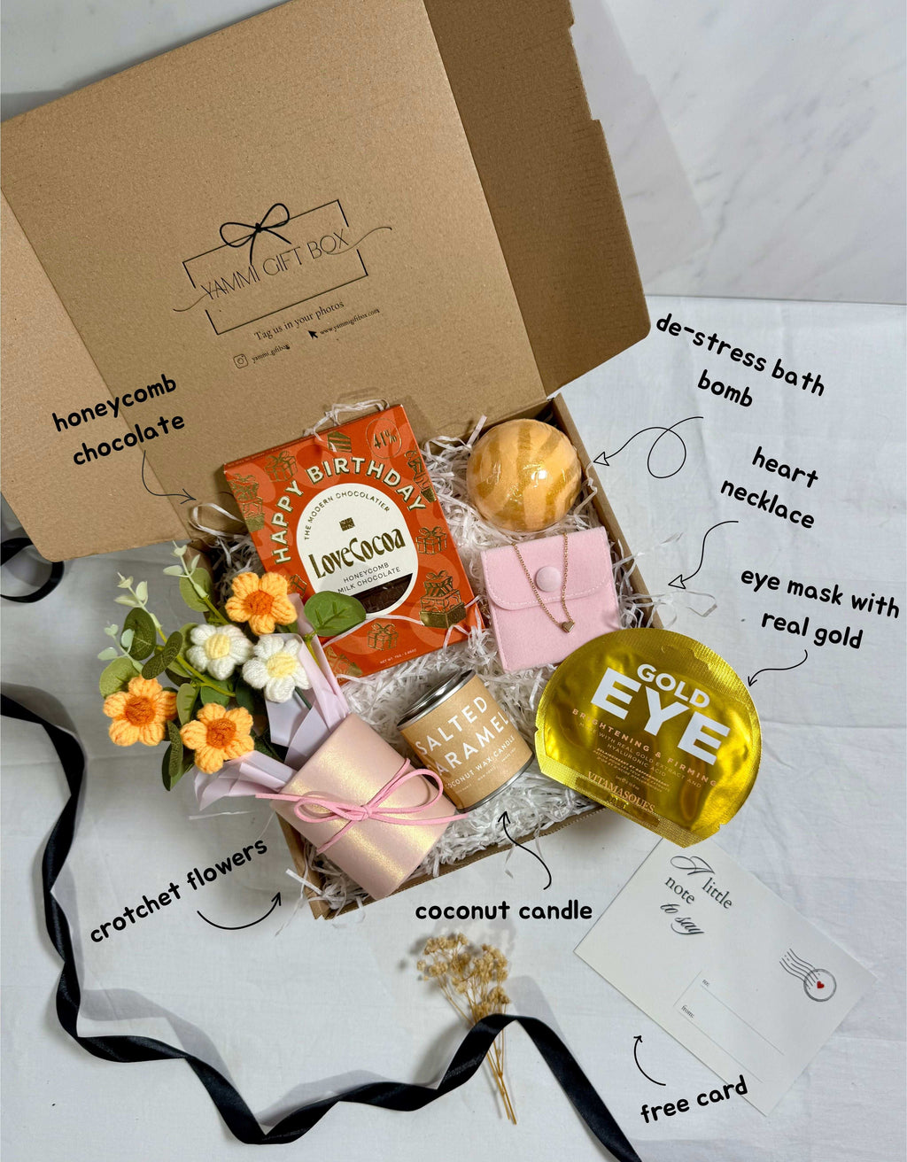 Gift box with various items including a gold eye mask, coconut candle, and flowers on a white background.