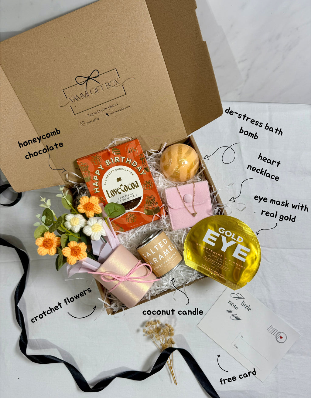 Gift box with various items including a gold eye mask, coconut candle, and flowers on a white background.