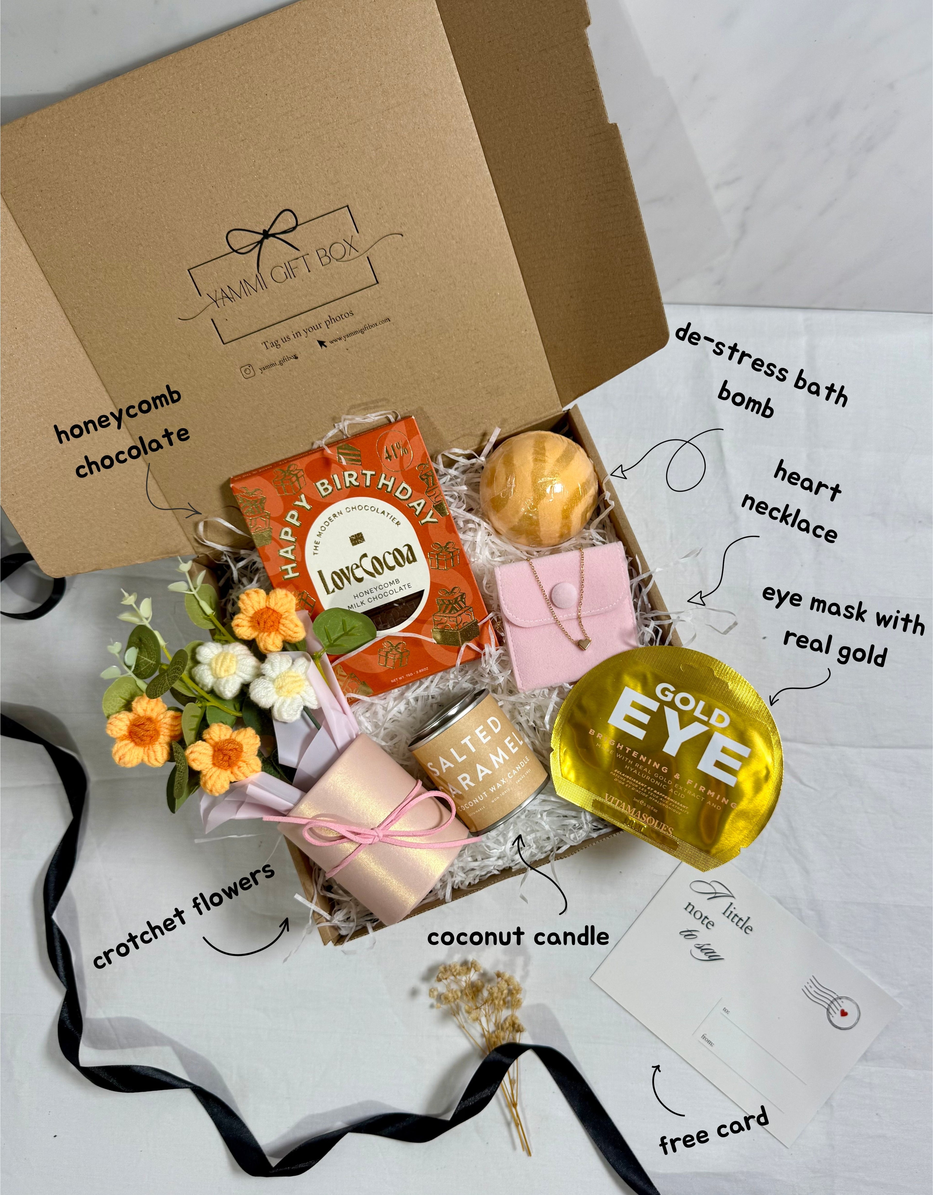 Gift box with various items including a gold eye mask, coconut candle, and flowers on a white background.