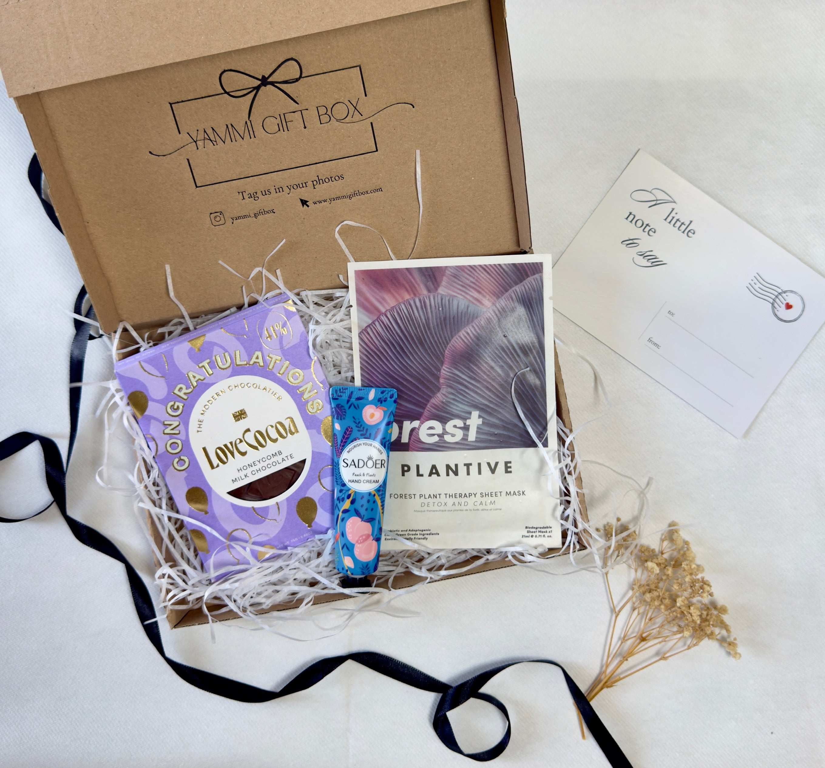 Congratulations gift box with purple Love Cocoa chocolate, Plantivia “Forest” sheet face mask, and blue floral Sadoer hand cream, styled with dried flowers, ribbon, and a “little note to say” card.