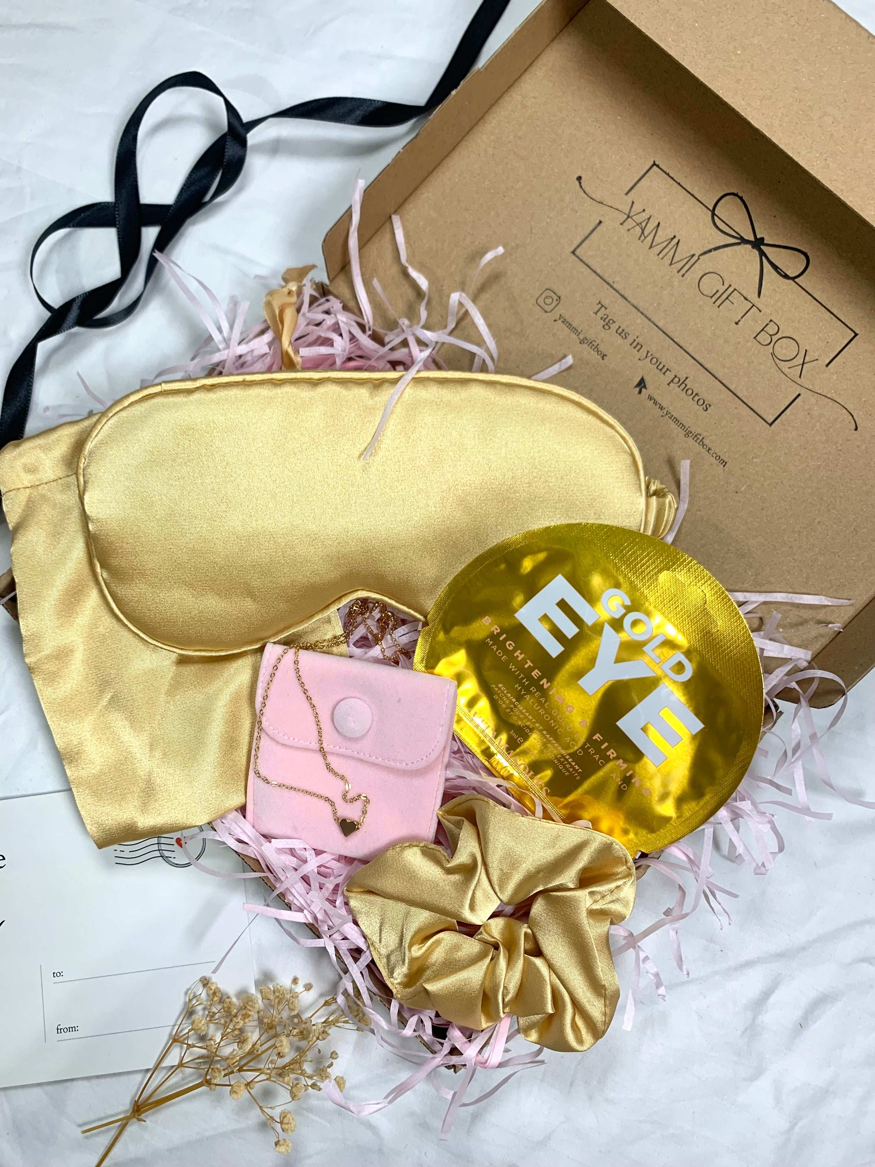 Luxury gold pamper gift box featuring satin eye mask, gold scrunchie, gold under-eye masks, pink pouch with gold heart bracelet, and dried flowers.