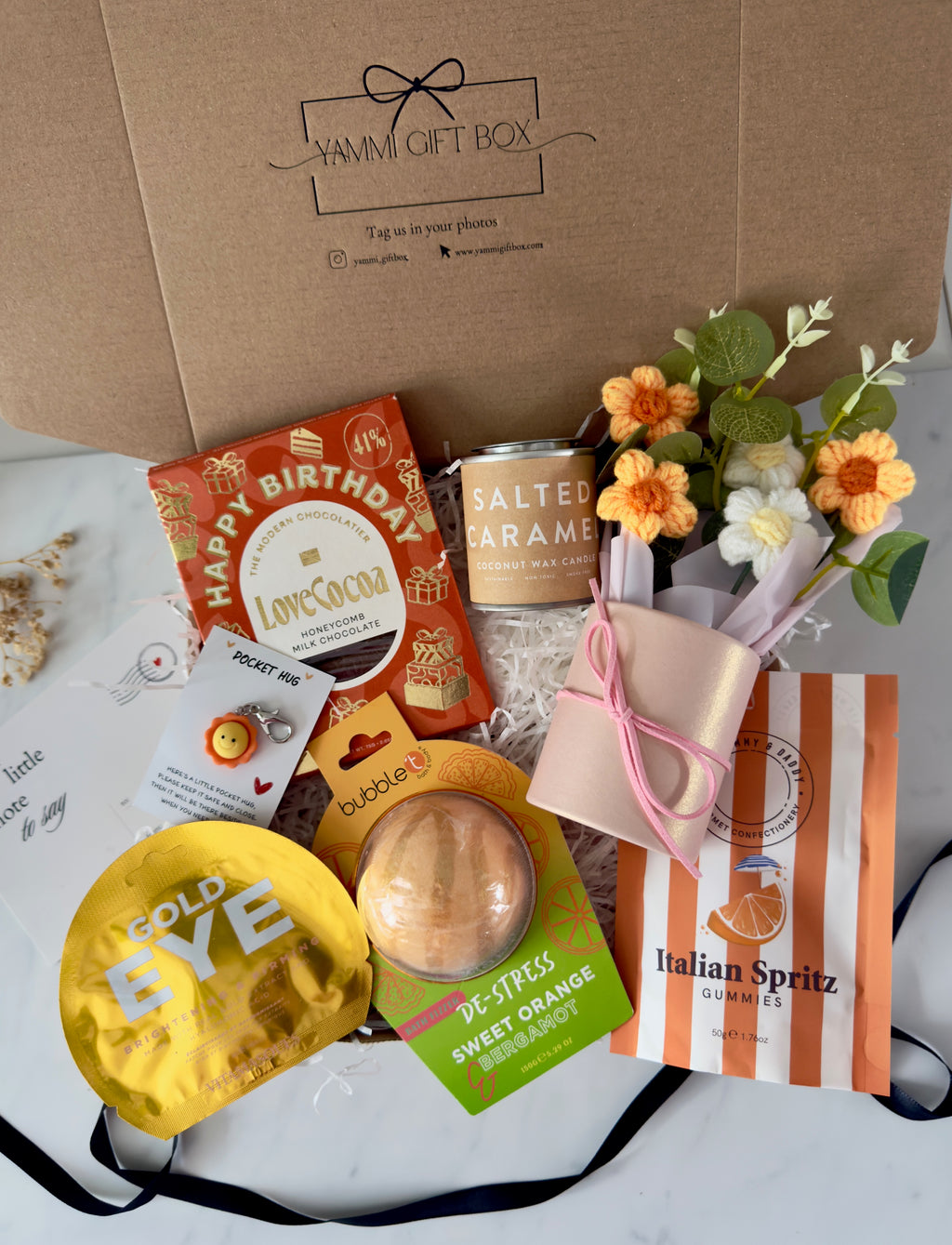 Birthday gift box for her with Happy Birthday chocolate bar, salted caramel candle, gold eye mask, sweet orange bath bomb, Italian Spritz gummies, pocket hug keyring, and crochet flower bouquet.