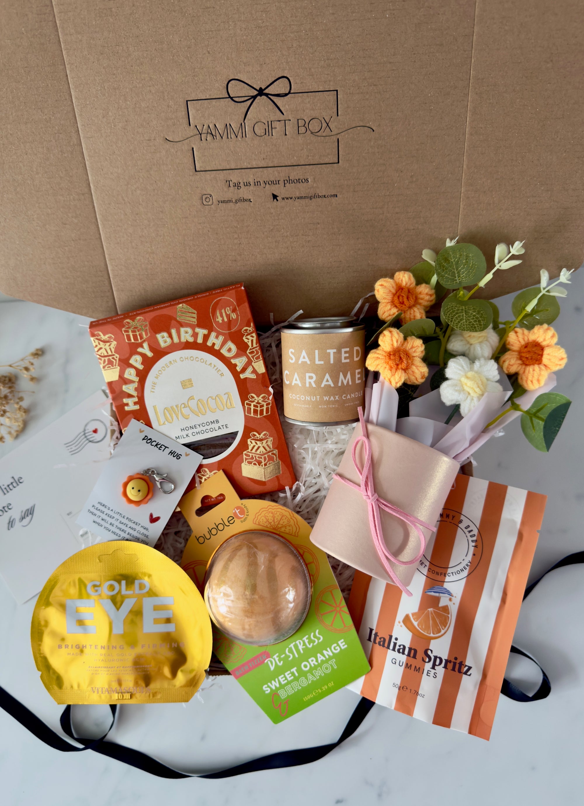 Luxury birthday gift box for her featuring Happy Birthday chocolate bar, salted caramel candle, gold under-eye mask, sweet orange bath bomb, Italian Spritz sweets, pocket hug token, and crochet flower bouquet.