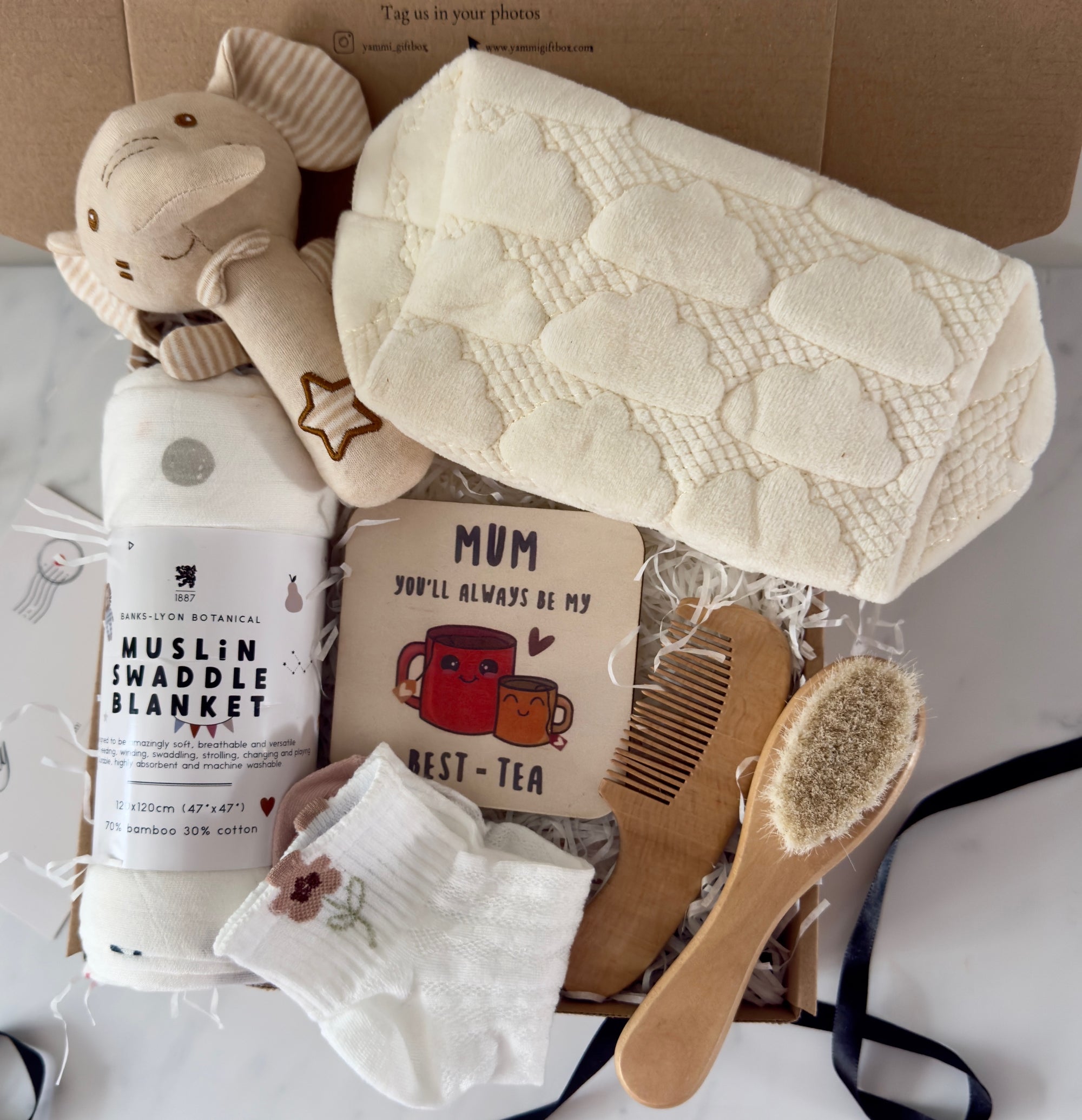 New Mum and Baby gift box including a soft muslin swaddle blanket, plush elephant toy, cream cloud-patterned knitted blanket, ‘Mum you’ll always be my best-tea’ keepsake card, baby hairbrush, wooden comb, and baby socks with floral embroidery – neatly presented in a Yammi Gift Box