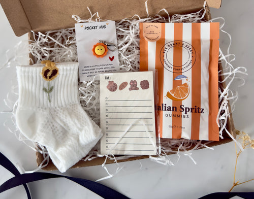 Close-up of Yammi Gift Box containing baby socks with embroidered sunflower, smiley face ‘Pocket Hug’ keyring, cute bear-themed notepad, and a striped pouch of Italian Spritz gummies — nestled in white shredded filler.