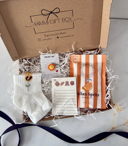 Yammi Gift Box featuring baby socks with embroidered sunflower, a smiley face ‘Pocket Hug’ pin, notepad with botanical design, and Italian Spritz gummies — presented in a kraft box with shredded white filler.