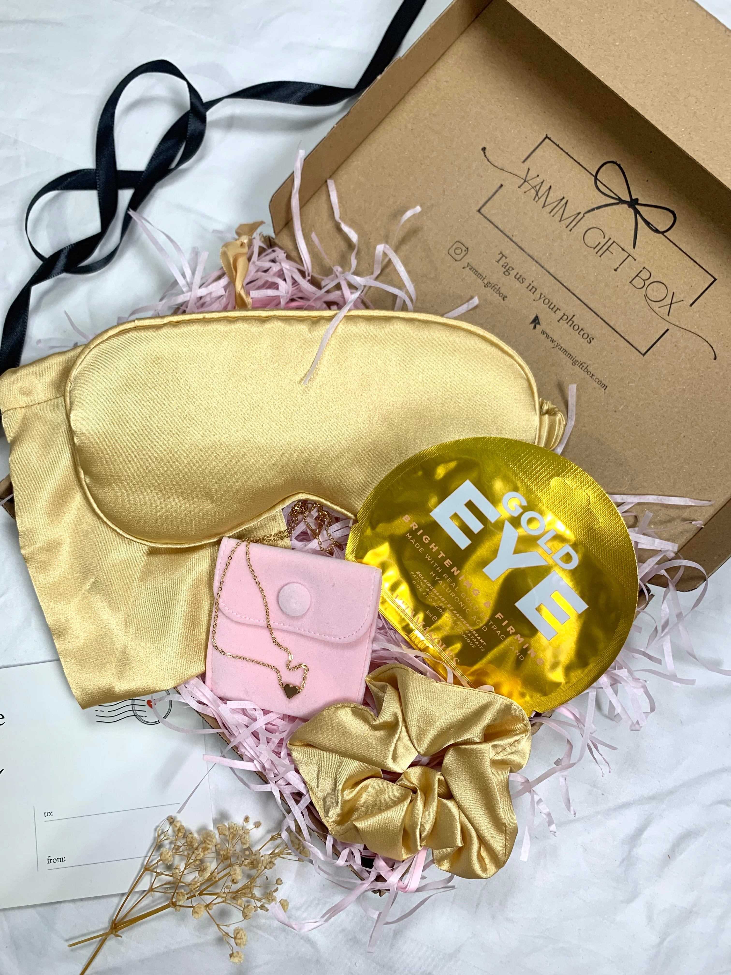 Luxury gold pamper gift box featuring satin eye mask, gold scrunchie, gold under-eye masks, pink pouch with gold heart bracelet, and dried flowers.