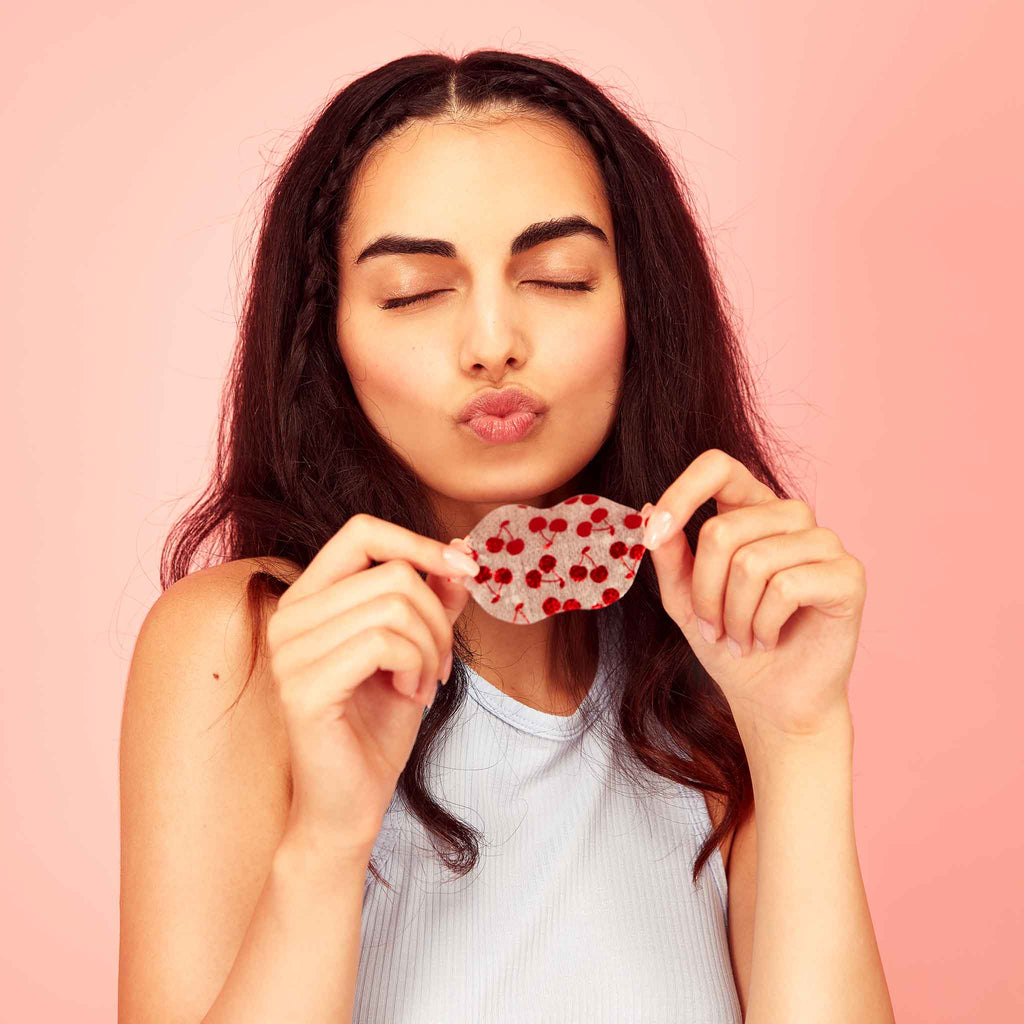 Woman holding cherry vegan collagen lip mask with red heart details against a pink background, making a kiss face. Hydrating, cruelty-free skincare for soft, plump lips.