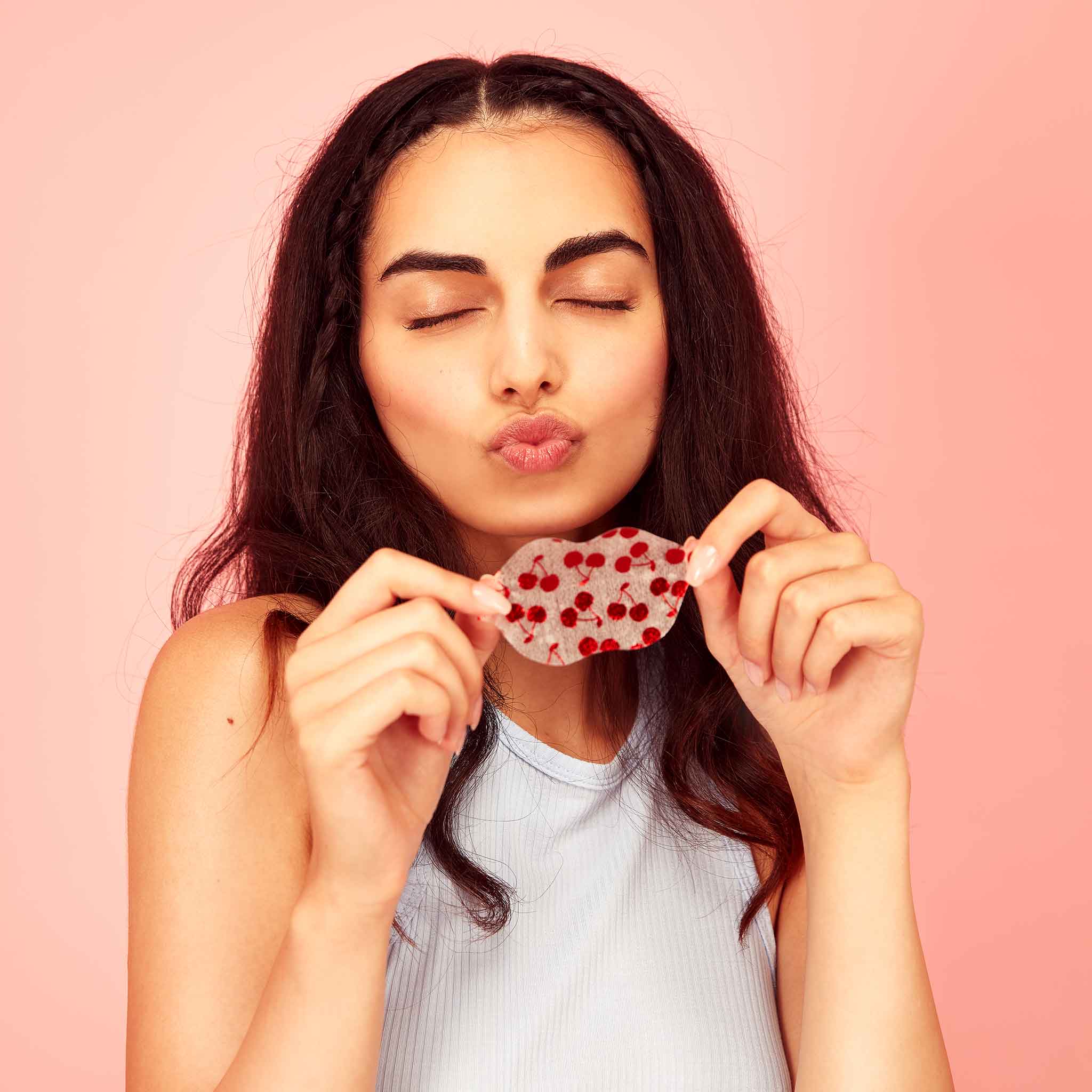 Woman holding cherry vegan collagen lip mask with red heart details against a pink background, making a kiss face. Hydrating, cruelty-free skincare for soft, plump lips.