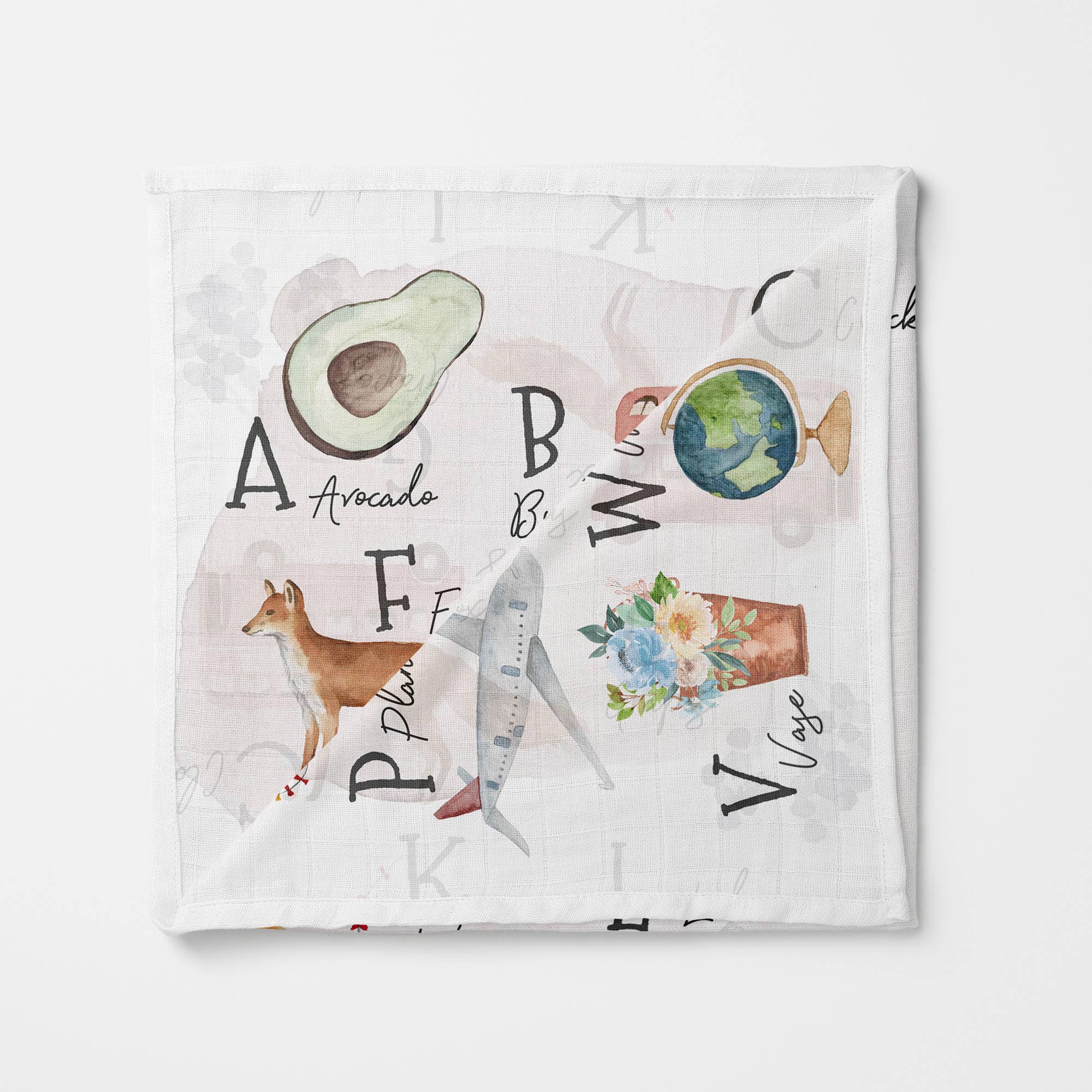 Organic baby muslin blanket with illustrated A–Z alphabet design, including watercolor drawings of an avocado, globe, fox, shark, flowers in a pot, and more — ideal for newborn gifts and educational nursery decor.