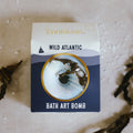 Navy and cream square box packaging for the Enniskinn Wild Atlantic Bath Art Bomb, featuring a circular transparent window that reveals the ocean-inspired bath bomb inside. The design includes minimalist wave graphics, a small sailboat icon, and bold typography. The brand name "Enniskinn" is printed in gold foil at the top, while "WILD ATLANTIC" and "BATH ART BOMB" are prominently displayed on the front, evoking a refreshing, coastal aesthetic.