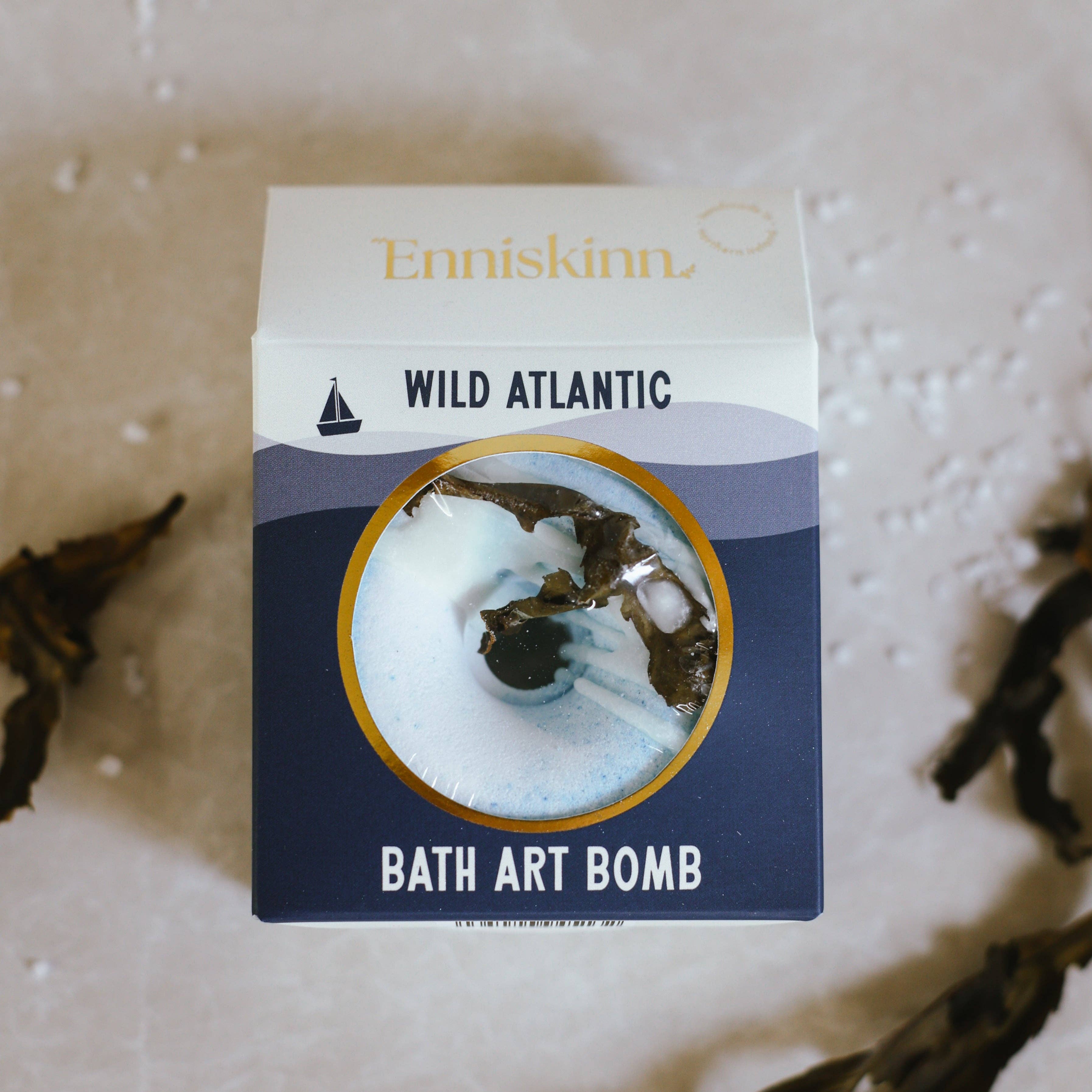 Navy and cream square box packaging for the Enniskinn Wild Atlantic Bath Art Bomb, featuring a circular transparent window that reveals the ocean-inspired bath bomb inside. The design includes minimalist wave graphics, a small sailboat icon, and bold typography. The brand name "Enniskinn" is printed in gold foil at the top, while "WILD ATLANTIC" and "BATH ART BOMB" are prominently displayed on the front, evoking a refreshing, coastal aesthetic.