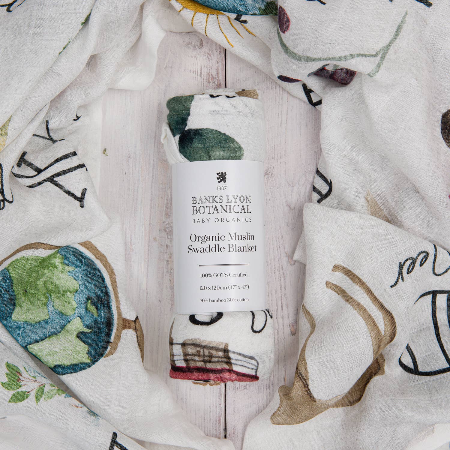 Organic muslin swaddle blanket by Banks Lyon Botanical, featuring a whimsical print with a globe, stacked vintage books, teacup and teapot, and leafy botanical illustrations — perfect for baby gifting and natural nursery themes.
