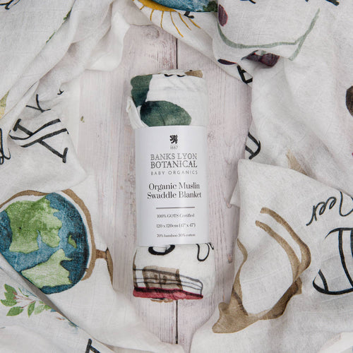 Organic muslin swaddle blanket by Banks Lyon Botanical, featuring a whimsical print with a globe, stacked vintage books, teacup and teapot, and leafy botanical illustrations — perfect for baby gifting and natural nursery themes.
